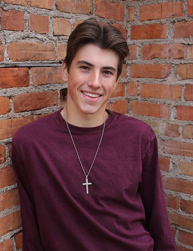 Blog author Cooper is standing in front of a brick wall. He has short brown hair and is wearing a purple long-sleeved shirt and a cross on a chain.