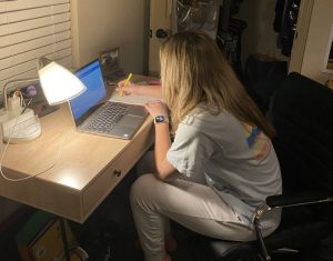 A young female college student with long blonde hair is sitting at student desk, looking at a laptop screen and taking notes.