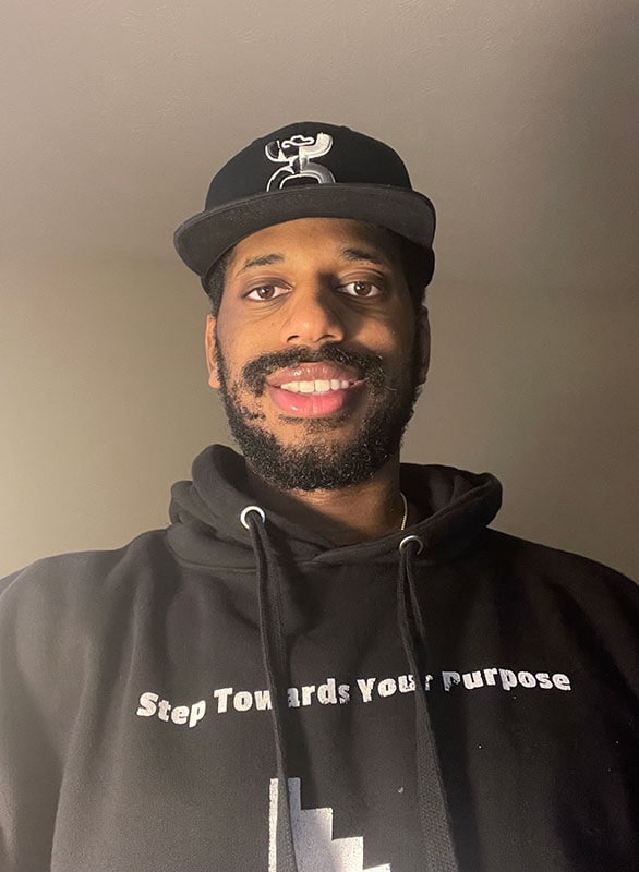 Blog author Chris Simmons is smiling and wearing a black baseball hat and a black hoodie with the words ‘Step Towards Your Purpose’ in white letters.