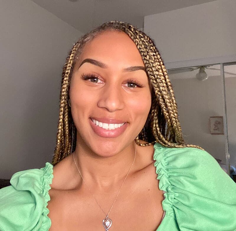 Blog author DaViane Lowe is smiling and wearing a bright green shirt ruffled at the shoulders and a silver heart pendant necklace. She has long light-colored braids.