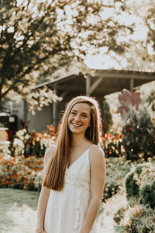 Blog author Emilie is standing outdoors in front of some flowering trees. She has long brown hair and is wearing a white dress.