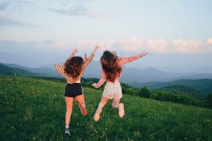 A mountain meadow with the Smoky Mountains in the distance. Two young women, with their backs to the viewer, are jumping up in the air.