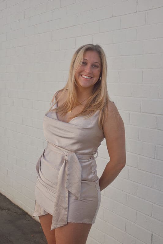 Blog author Emma Karant is smiling and standing in front of a white brick wall. She has medium length blonde hair and is wearing a white dress with a sash at the waist.