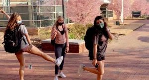 Three female college students stand outside on their campus with their backpacks. They are wearing facemasks.