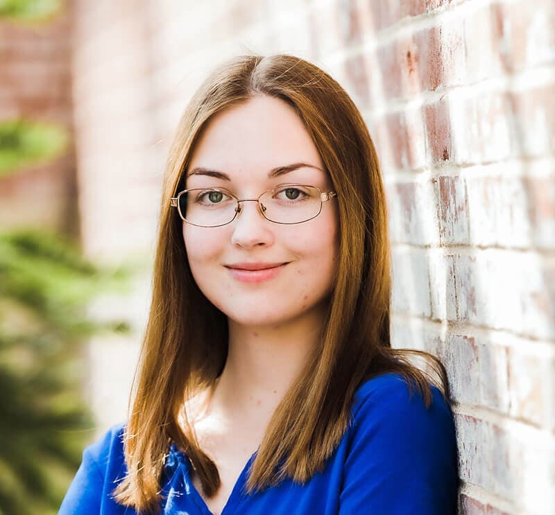 Blog author Faith is smiling and leaning against a brick wall. She has straight brown hair and is wearing wire-rimmed glasses. 