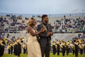 A female and male student are standing arm-in-arm on a football field during homecoming. The female is wearing a long gold gown. The male is wearing a dark suit with a black shirt. They each wear a sash that says, “Homecoming Court”.