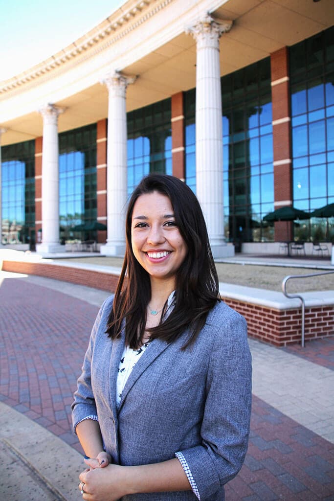 Blog author Jasmine stands outside in front of an academic building with white pillars. She has medium length dark hair and is wearing a light gray blazer with a white blouse.