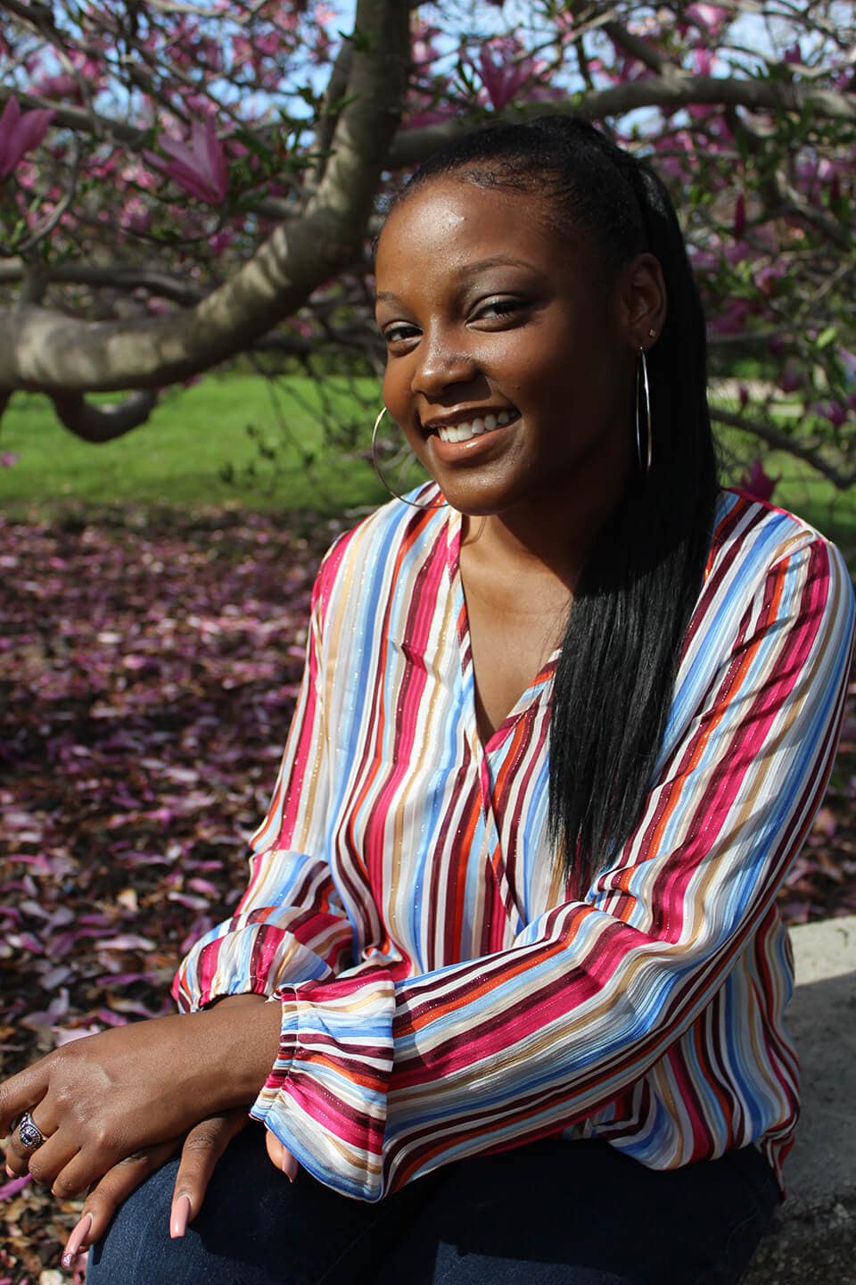 Blog author Jayla Pope is outside standing in front of a tree. Her long black hair is in a ponytail and she is wearing large silver hoop earrings and a colorful striped blouse.