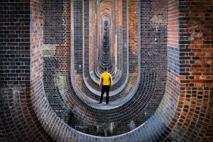 A man wearing black jeans and a yellow jacket stands with his back to the viewer, looking down in a series of brick archways.