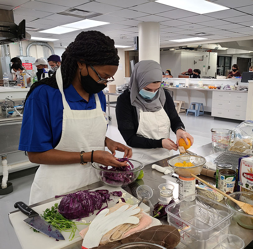 Two college students are wearing face masks and aprons and prepping food in an industrial-type kitchen. The student on the left is chopping cabbage. The student on the right is zesting a lemon.