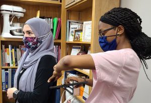 Two college students standing in front of a bookcase with cookbooks and cooking equipment. They are both wearing face masks and the student on the left is also wearing a headscarf.