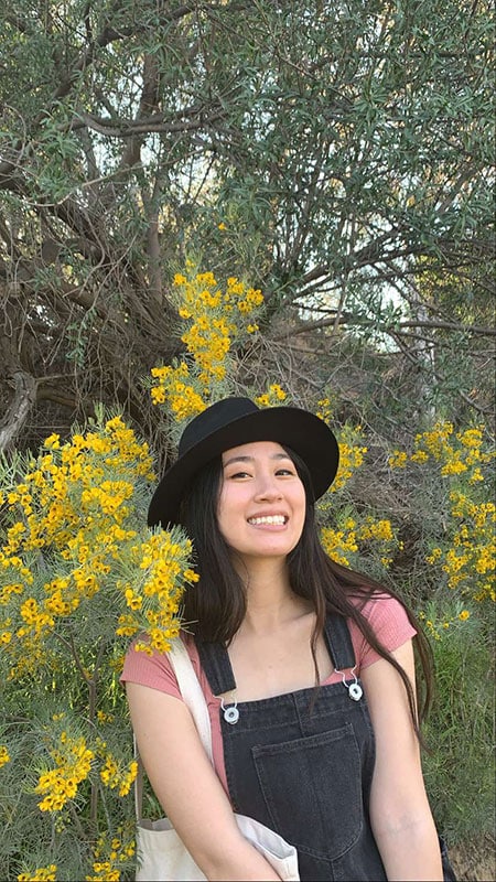 Blog author Kaitlin Hung is standing outside in front of a tree with yellow leaves. She has long black hair and is wearing a black hat and black tank top with a white shoulder bag on her right shoulder.