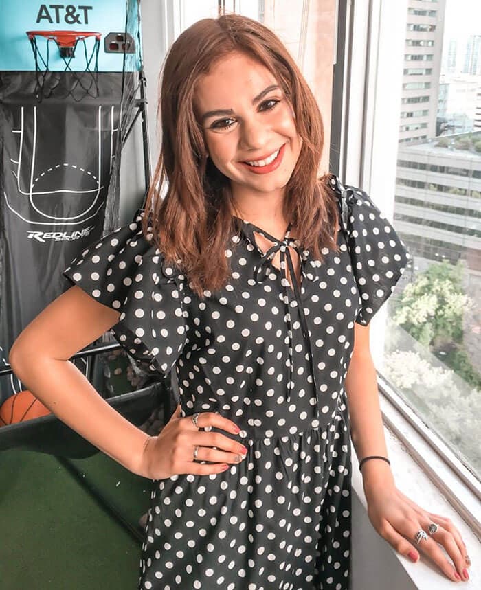 Blog author Kiara Lozano is smiling and standing in front of an open window. She is wearing a black blouse with white polka dots and has her hand on her hip.