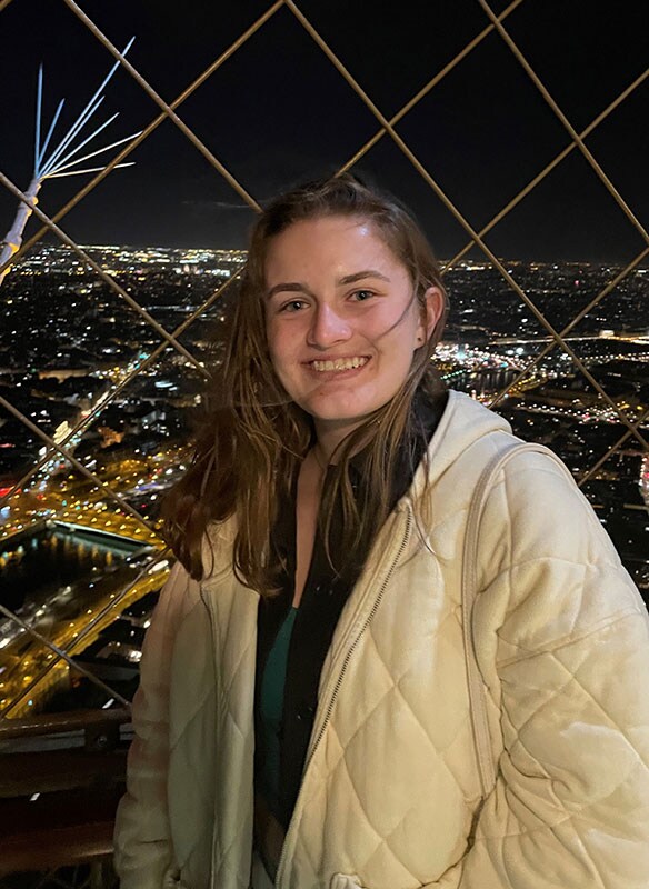 Blog author Kennedy is standing on a bridge at night with the lights of a city behind her. She has long brown hair and is wearing a light-colored winter coat.