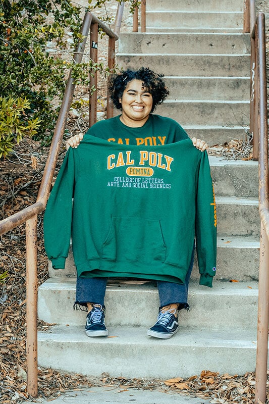 Blog author Katie is sitting on concrete steps and hold up a green Cal Poly Pomona sweatshirt.