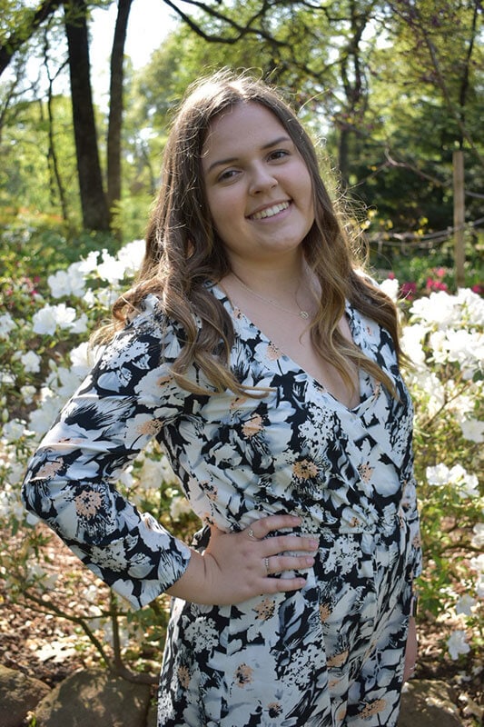 Blog author Kara Stevens is smiling and wearing a black floral dress and standing outside with trees in the background. She has shoulder length brown hair.