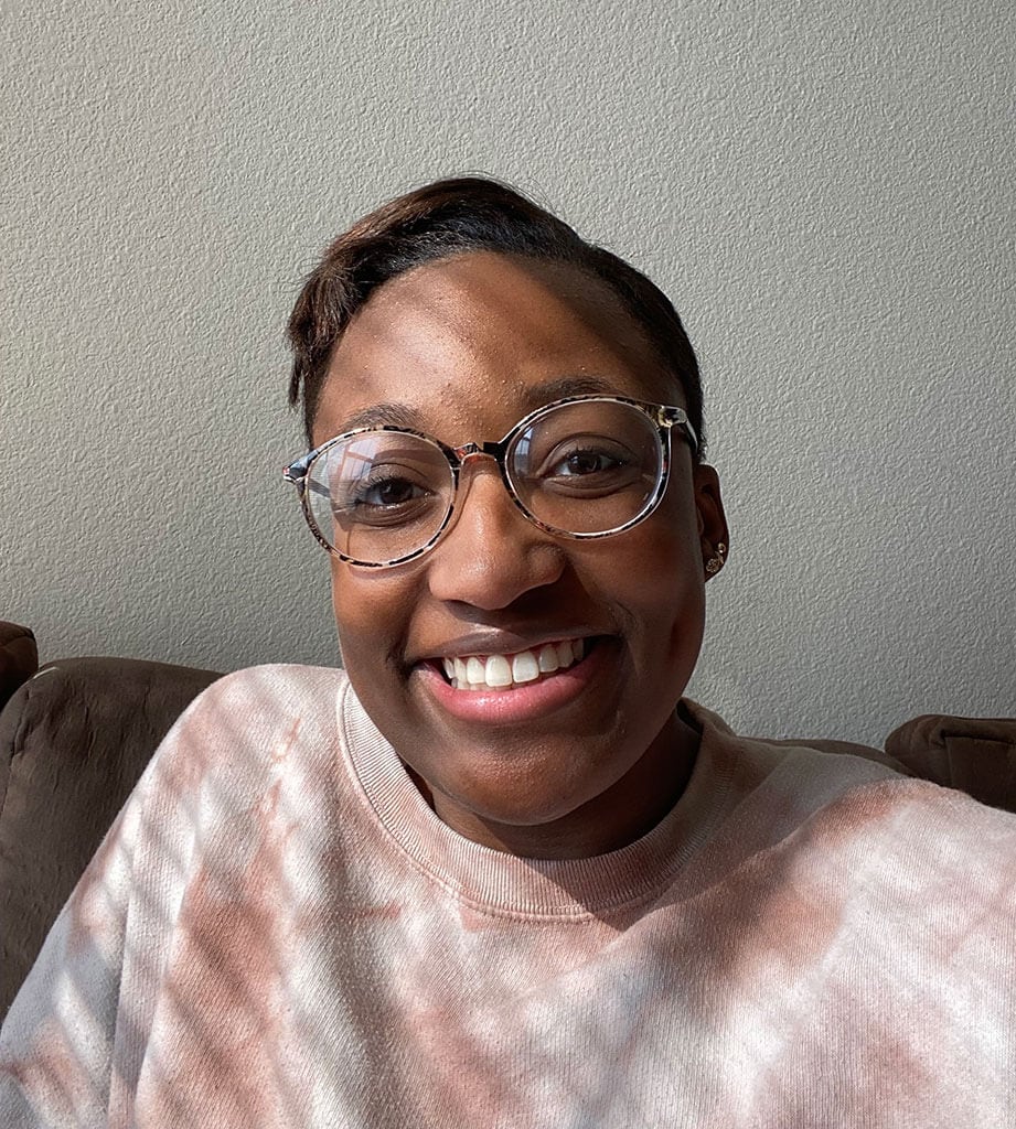 Blog author KC Cooper has short, dark hair and wears round glasses. She is sitting on a brown couch and wears a white sweatshirt with a floral print.