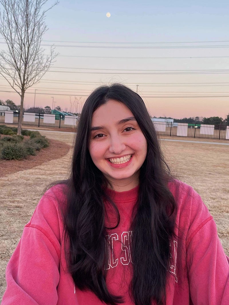 Blog author Laura Avellaneda stands outside with the sun setting behind her. She has long brown hair parted in the middle. She is smiling and wearing a red sweatshirt.