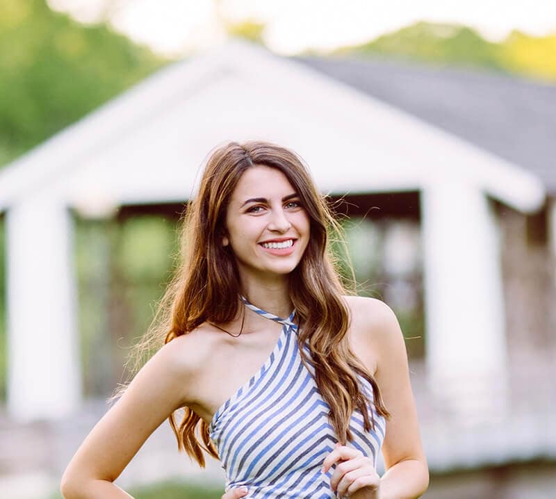 Blog author Logan Collins stands outside in front of a white building. She has long brown hair and is wearing a blue and white striped halter top.