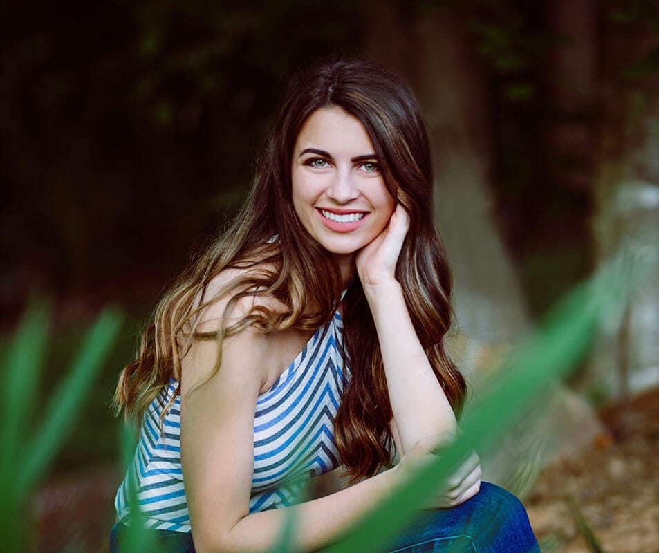 Blog author Logan Collins sits outside with trees in the background. She has long brown hair and is wearing a sleeveless blue and white striped shirt. 