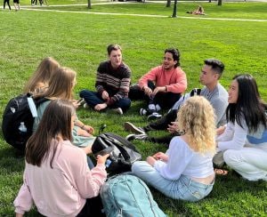 A group of male and female college students sitting in a circle and talking on a campus lawn. Their backpacks are scattered on the grass beside each student.