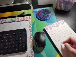 A view looking down on a college student’s desk featuring a large desk calendar, computer keyboard, computer mouse, and notepad. The student’s hand appears to be writing notes on the notepad.