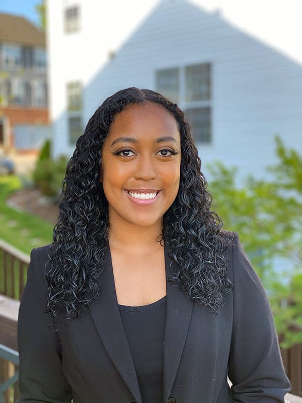 blog author Mariam Ameha is standing outside with a white 2-story house behind her. She has dark curly hair and is wearing a business suit jacket and dark top.