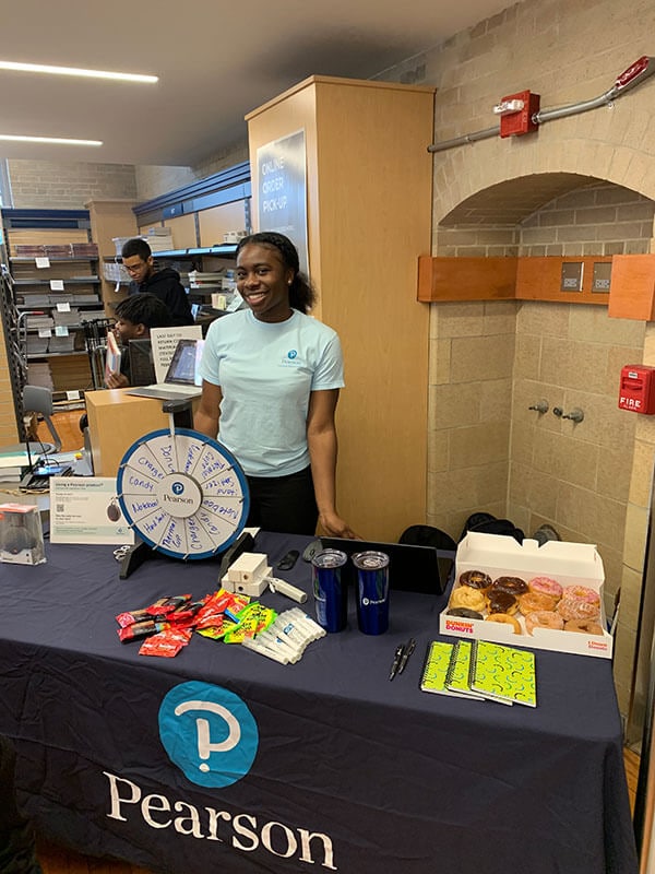 Blog author Mercy Aruleba, a young female college student, is standing behind a table with promo items and a box of donuts. She is working in her capacity as Pearson Campus Ambassador.