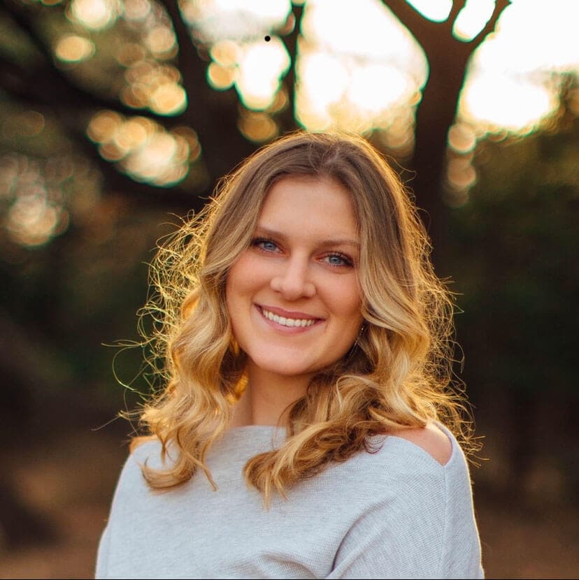 Blog author Madison Butler is standing outside in front of green trees. She has long blond wavy hair and is wearing a light grey long sleeved sweater.