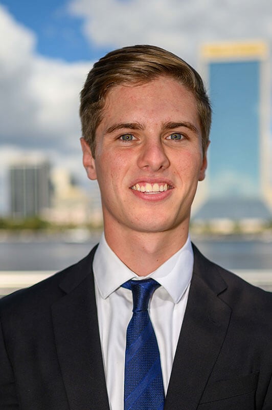 Blog author Matthew Dougherty is smiling with a blurred view of a city behind him. He has short light brown hair and is wearing a dark suit jacket, white shirt, and dark blue tie.
