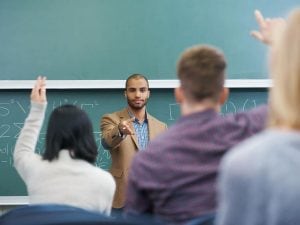 A young Black college professor stands in front of the classroom with his back to a green chalkboard. Several students seated in front of him are raising their hands.