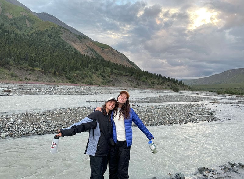 Blog author Maeve and a friend are standing are-in-arm near a mountain stream in Alaska.