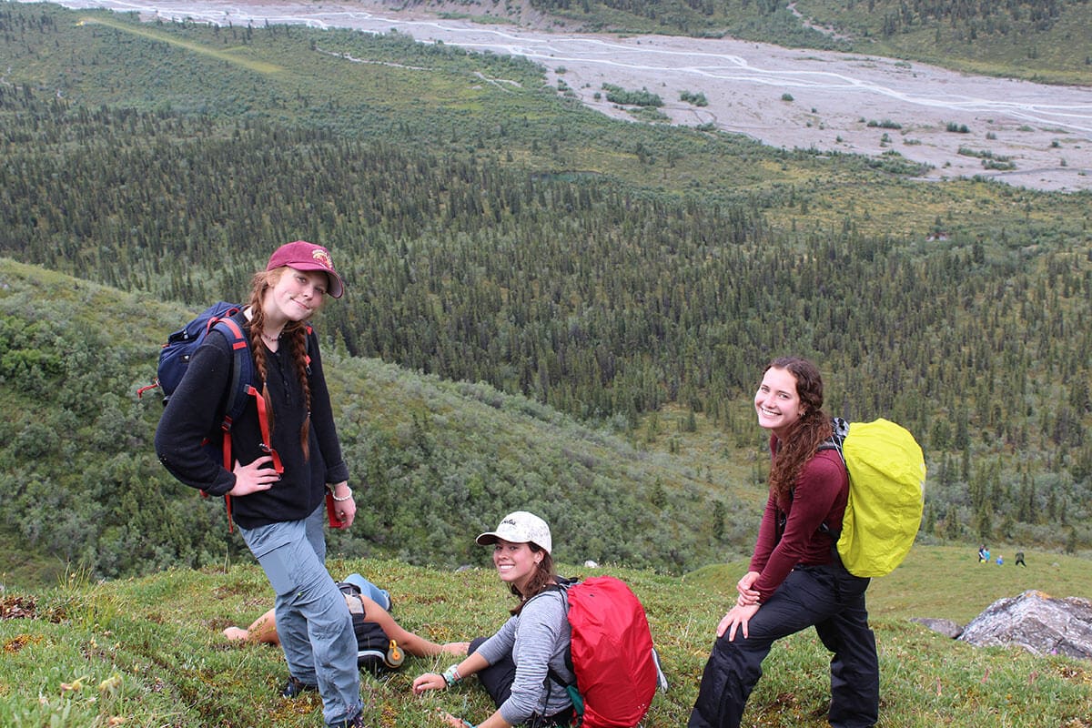 Blog author Maeve and two friends are standing on a steep hill wearing backpacks and looking up towards the camera. There is a mountain stream below at the foot of the hill.