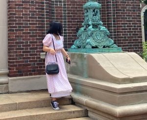 Blog author Miyu Nakajima is wearing one of her vintage dresses, a long pink dress with short, puffed sleeves and belted at the waist. She is also wearing black Converse sneakers and has a black cross-body purse. She is standing on concrete steps and is looking back towards something behind her.