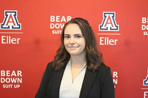 Blog author Mikayla is standing in front of a red wall with logos from the University of Arizona Eller School of Business. She has medium length brown hair and is wearing a navy blazer over a white shirt. 