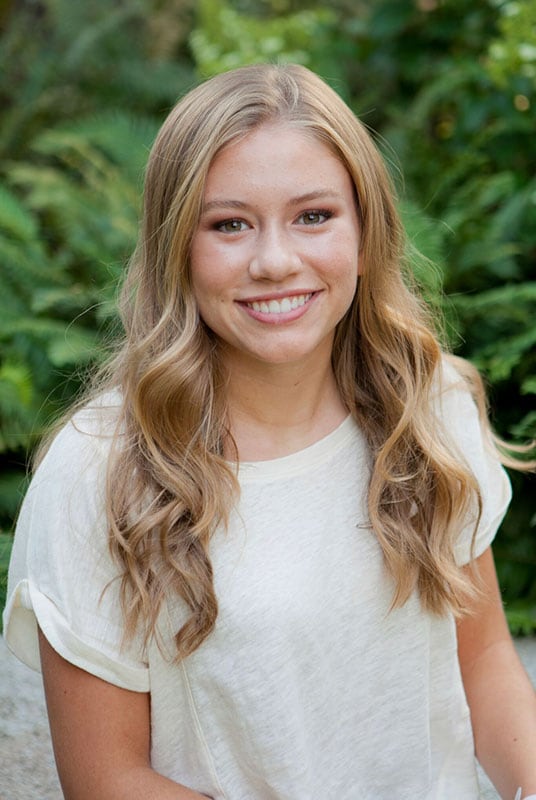 Blog author Olivia Kane is smiling and standing in front of a leafy green background. She has medium length blond hair and is wearing a white short-sleeved shirt.