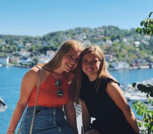 Two female college students sitting side-by-side with a lake and shoreline in the background. Location: Arendal, Norway