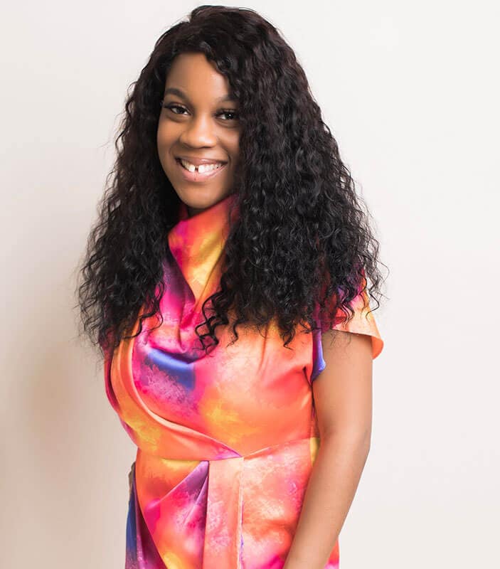 Blog author Princess Robinson is smiling and standing in front of a bright white wall. She has long black curly hair and is wearing a brightly colored short-sleeved dress.