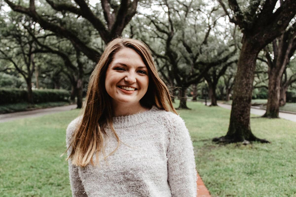 Blog author Rachel Calcote is smiling and wearing a cream-colored sweater. She has medium length brown hair. She is standing outside in front of a grove of large trees.