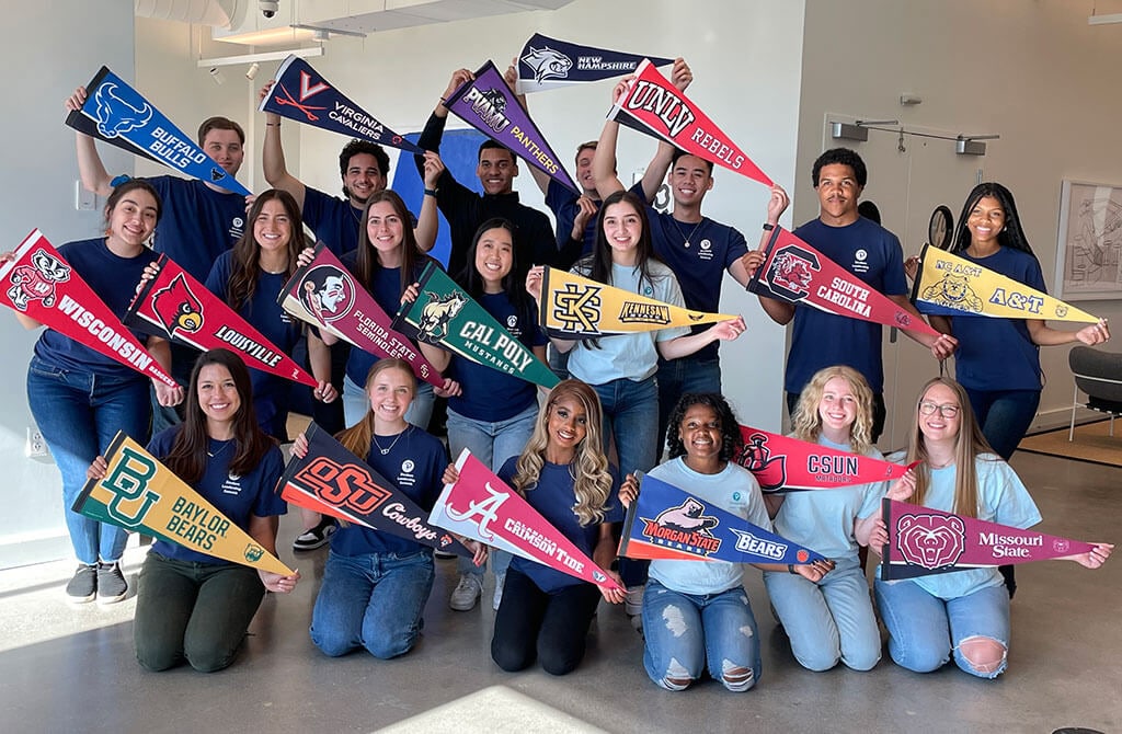 A group of 18 college students are standing close together in an office setting. Each student is holding a college pennant from their school. 