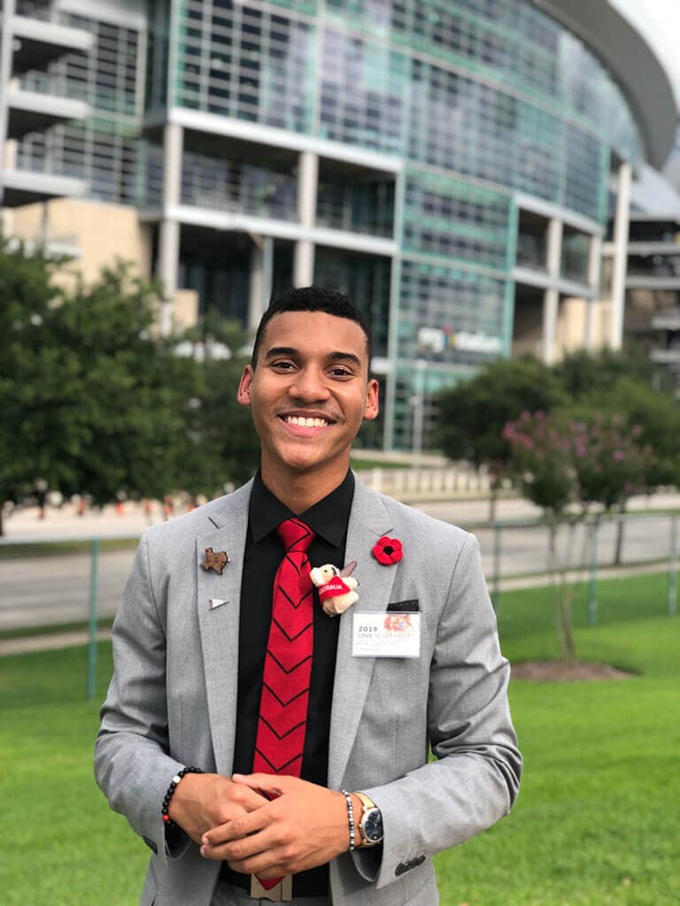 Blog author Ryan Celestine stands outside in front of a building on his campus. He is wearing a grey suit, black shirt and red necktie.