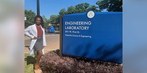 A woman wearing a white lab coat standing beside a college campus sign that reads, ‘Engineering Laboratory’.
