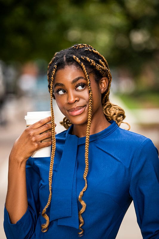 Blog author Synia Malbrough is pictured wearing a blue button shirt and holding a to-go coffee. She has long braids and is looking up to her left.