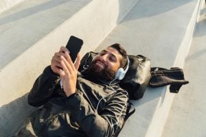 An adult male with dark hair and beard is lying on his back with his head on a backpack. He is wearing headphones and looking at his mobile phone.
