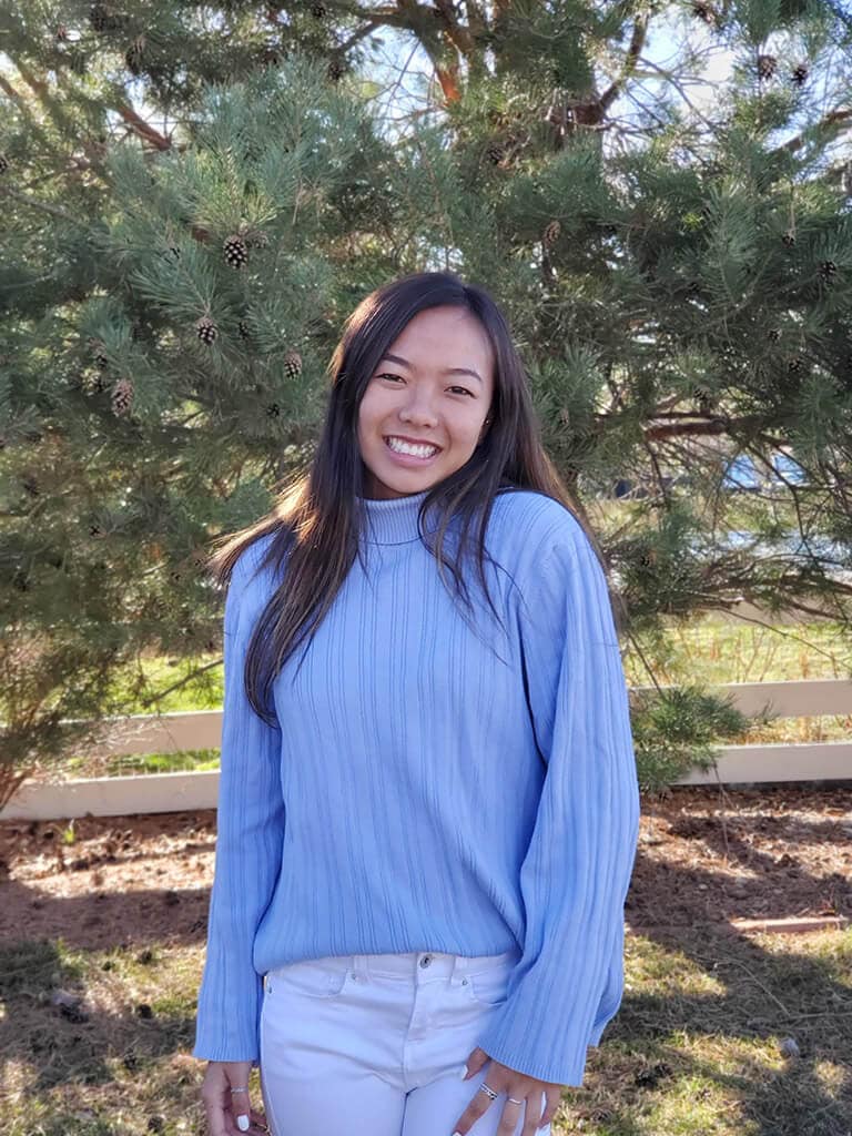 Blog author Sydnie Ho is standing outside in front of some trees. She has long black hair and is wearing a blue long-sleeved sweater.