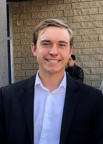 Blog author Tristan Larking is smiling and standing in front of a stone wall. He has short brown hair and is wearing a navy blue blazer and light blue shirt.