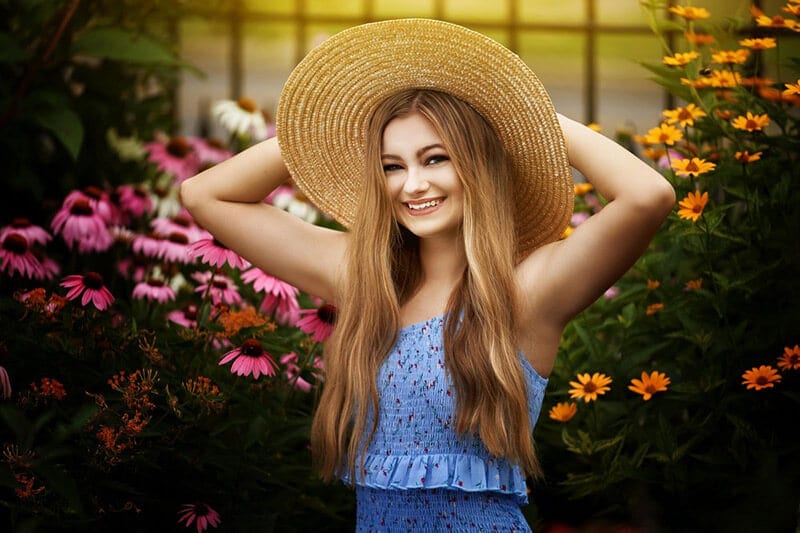 Blog author Taylor Perline is smiling and standing in front of a batch of blue and yellow flowers. She is wearing a brown straw hat with a large brim and a blue dress.