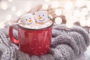 A red mug of hot chocolate set on top of a gray knit blanket. The hot chocolate is topped with two marshmallows with snowmen faces. 