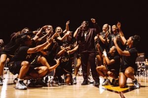A group of college women basketball players in black uniforms crowded around a coach speaking into a microphone.