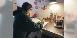 A college student sits at a student desk writing in a notebook. There are various student office supplies on the desk. The student is wearing noise-canceling headphones.
